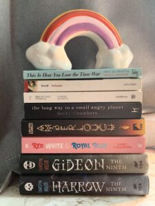 A pile of books with a rainbow sculpture on top. The titles are, from top to bottom: This is how you lose the time war by Amal El Mohtar and Max Gladstone, Orlando by Virgina Woolf, Camere Separate by Pier Vittorio Tondelli, The long way to a small angry planet by Beeky Chambers, Amberlough by Lara Elena Donnelly, Red, White and Royal Blue by Casey McQuiston, Gideon The Ninth by Tamsyn Muir and Harrow The Ninth by Tamsyn Muir.
