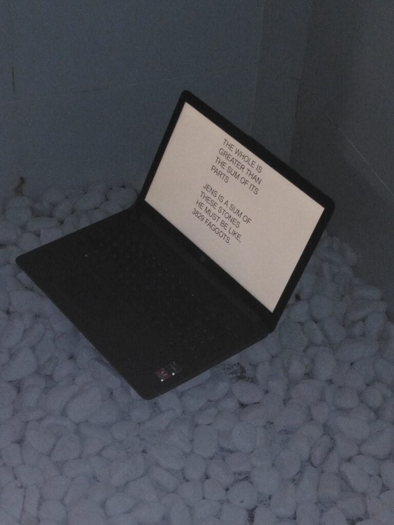 image of a shower filled with small rocks. In the middle standing a 13-inch laptop, reading text. The laptop shows a white screen with black letters filling the screen, reading: "THE WHOLE IS GREATER THAN THE SUM OF ITS PARTS JENS IS A SUM OF THESE STONES HE MUST BE LIKE, 3829 FAGGOTS"