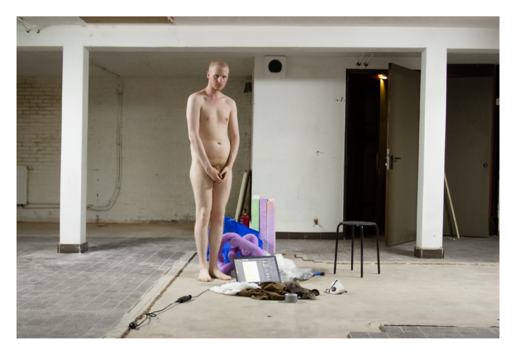 A picture of a bald, white person who is assigned male at birth standing nude in the middle of a hallway, made of concrete bricks painted white. The person covers their genitals. Right to them stands a black chair, on the floor right to them lie a laptop, brown and white fabrics, gray tape, a white mouthpiece, transparant plastics, blue plastics, a fluffy purple structure and green/purple bricks.